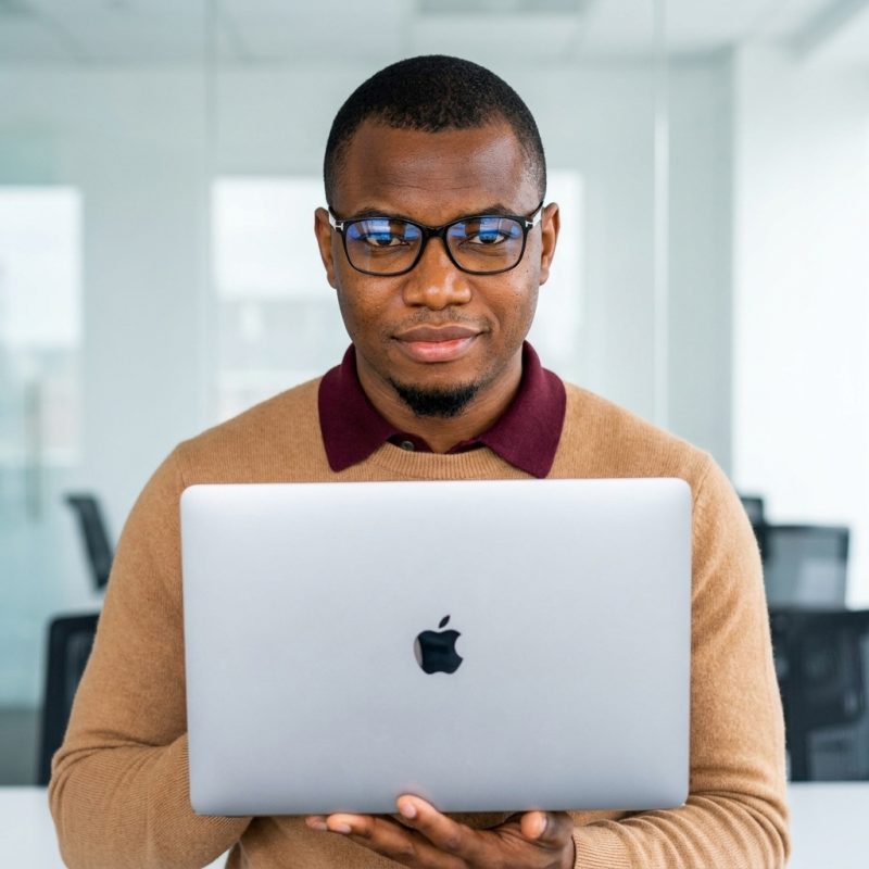 Jeune entrepreneur africain à lunettes tenant un MacBook dans un bureau moderne lumineux